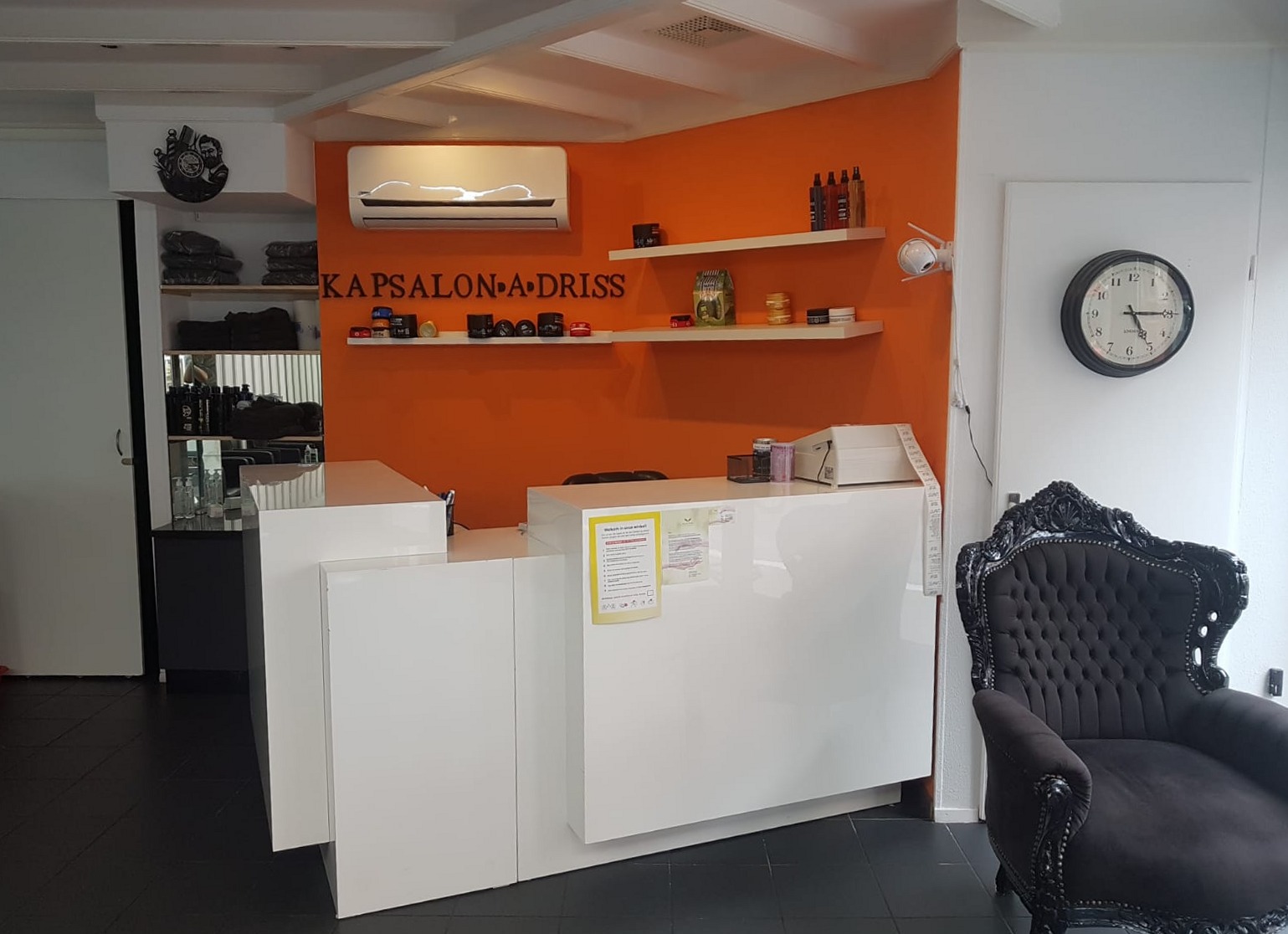 De balie van Kapsalon Driss voor de oranje wand met de naam van de zaak, met rechts een zwarte barokstoel