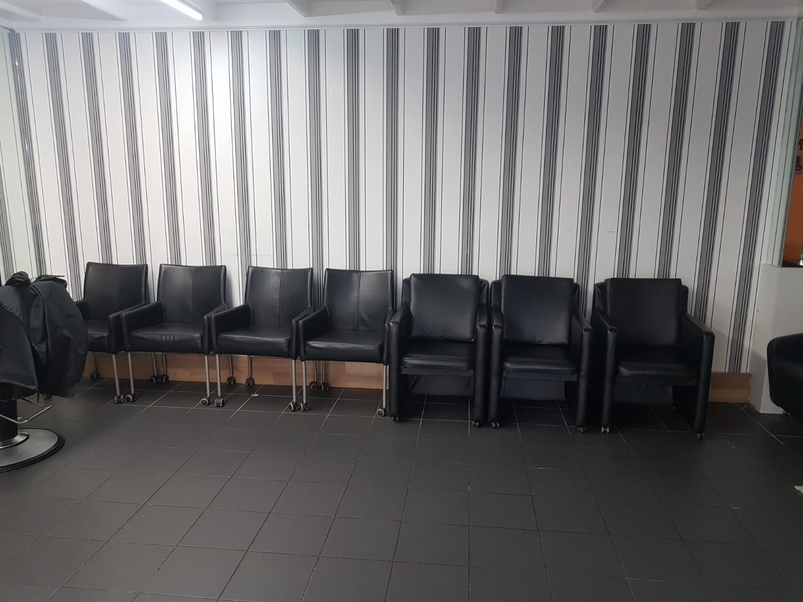 Een rij zwarte wachtstoelen voor de gestreepte wand in Kapsalon Driss